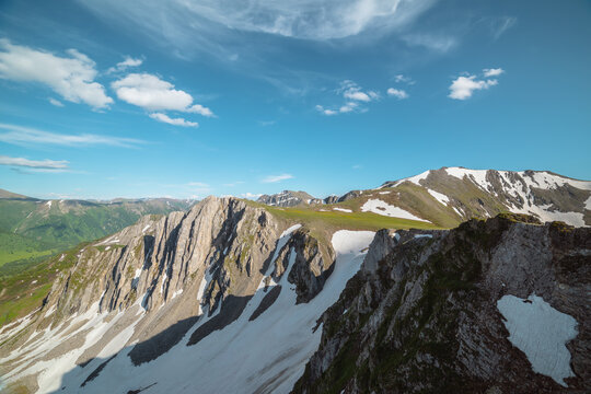 Aerial dizzying view to rocky cliff among sharp rocks under clouds in blue sky. Vertigo scenery with sheer crags above precipice, snowy ridge and mountain top far away in cloudy changeable weather. - Powered by Adobe