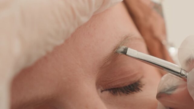 closeup eyebrow microblading on closed eye, esthetician applies pigment with fine tool wearing sterile gloves, soft neutral lighting, relaxed client, subtle contour enhancement, detailed precise