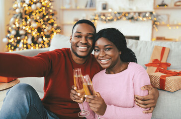 Obraz premium Two individuals smile and hold glasses of drink while sitting on a couch in a festive living room. There are holiday decorations and wrapped gifts nearby, creating a joyful scene.