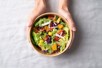 Healthy vegetable salad with lettuce, tomato, pumpkin, corn, purple sweet potato, cabbage, azuki bean and crab stick in takeout bowl with hand, Top view
