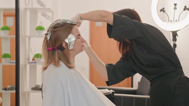 Stylist applying foils to hair, closeup salon color session with careful foil placement, ring light glow, cape and clips, focused hands, soft natural lighting, modern interior, precise technique,