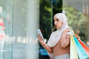 Huge discounts, great sale, message in app, cashback for buyer and device for shopaholic. Shocked cute young islamic woman in hijab with colored shopping bags typing on smartphone near shop window