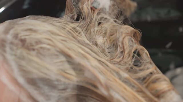 Stylist rinsing and detangling blonde hair with sudsy treatment, closeup of hands pulling through wet strands to remove product, visible sink basin and towel, professional salon workflow and crisp
