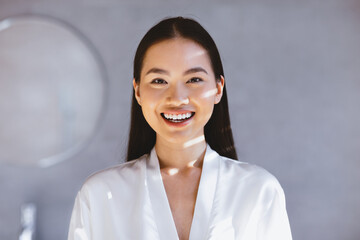 A woman smiles while wearing a white robe in a well-lit indoor room. Soft light filters through the...