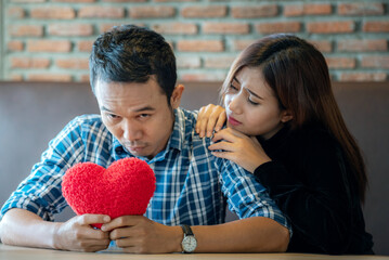 A man and a woman are sitting at a table. The man is holding a red heart-shaped pillow and looking sad. The woman is sitting next to him with her hand on his shoulder. She is looking at him with conce