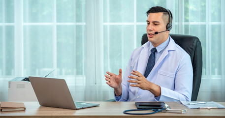 Fototapeta premium Confident male doctor in headset having video call on laptop in modern clinic