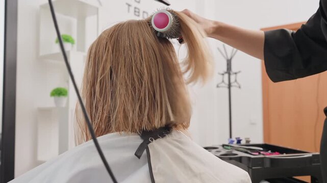 Curly bob receiving professional blowout with dryer, stylist uses round brush and high speed dryer to smooth damp bleached curls, bright modern salon, white cape, product trolley and ring light, quick