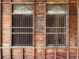 Traditional wooden house facade featuring two vintage Thai-style windows with ornate carved ventilation panels. Natural aged timber texture, rustic architecture, cultural craftsmanship, and classic 