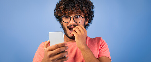 A man with curly hair is surprised while looking at his phone. He is biting his nails and has a shocked expression on his face. The blue background adds color to the scene.