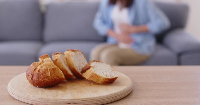 Gluten bread slices lying on wooden board, trigger for food intolerance and celiac disease in woman. Girl suffering from allergic symptoms, stomach pain and indigestion, holding belly, sitting on sofa