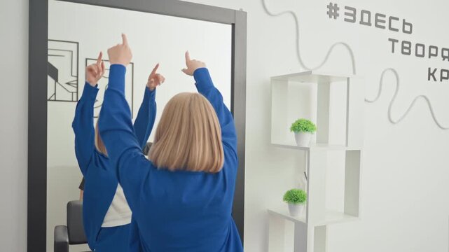 caucasian woman tries mirror makeover during salon consultation, stylist adjusts hair and gives guidance, blue jacket in white creative studio with shelves and plants, playful gestures evolve into