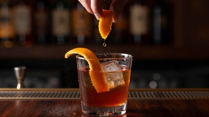 A hand garnishes a dark cocktail in a glass with an orange peel and ice cubes