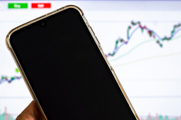 Man's hand holding a smartphone in front of a screen with a stock market chart, symbolizing mobile trading, investment, and financial analysis in the digital age.