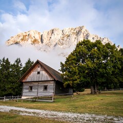 A rustic wooden cabin rests below a majestic mountain peak enveloped in clouds. Lush green trees surround the serene landscape