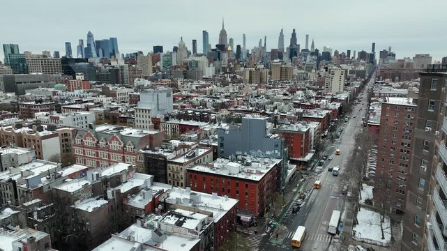 New York City showing snow-dusted rooftops, dense residential blocks and traffic along wide urban avenue under cold overcast sky. Yellow school buses on street. Aerial Rising wide shot.