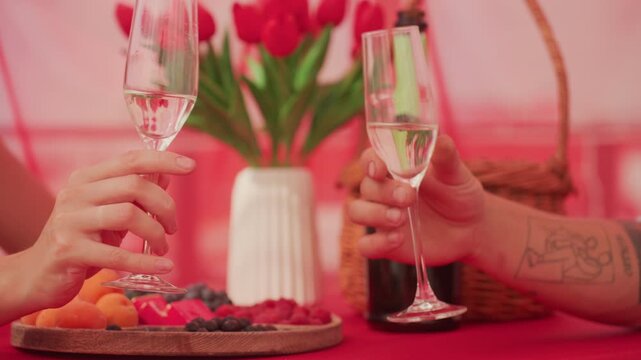 two champagne glasses on picnic table, hands clinking over platter of berries and apricots, tulip bouquet in ceramic vase, wicker basket and champagne bottle, soft pink lighting and tattooed arm