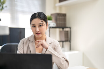 Pretty asian businesswoman or office worker putting a hand under chin and looking at laptop on table