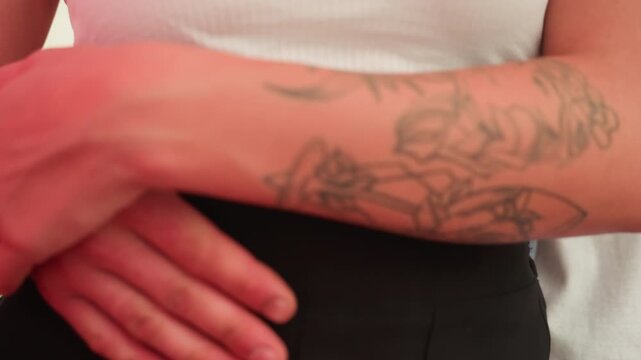 tattooed arms embrace waist closely, tender date moment, hands clasped over black pants, white crop top visible, soft red lighting, close framing highlights floral forearm tattoo, gentle squeeze
