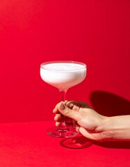 Hand Holding a Creamy White Cocktail in a Coupe Glass Against a Vibrant Red Background with Dramatic Shadows