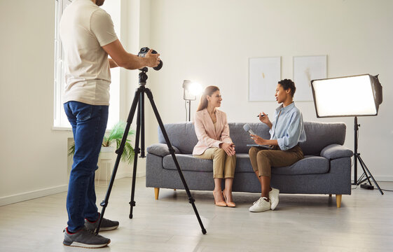 A camera operator recordong video interview in a bright studio with two hosts seated on a couch under production lights and tripod. Woman journalist with microphone interviewing young guest