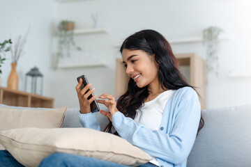 Smiling pretty young Asian woman sitting on cozy couch, using funny mobile apps in living room. Woman at Home, Doing Online Shopping, Messaging Friends, Posting on Social Media