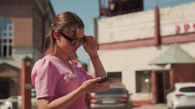 young woman in pink dress checking phone and adjusting sunglasses, storefront and parked cars behind, candid midshot showing concentration on messages and navigation gestures, sunlit street atmosphere