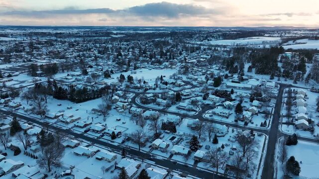 Rural american landscape with snowy neighborhood in winter season. Aerial backwards wide shot. Blue colored scenery at sunrise. Row of houses covered in snow and ice. Straight street cleared from snow