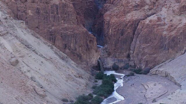 Drone shot of a muddy flash flood waterfall cascading into the deep Kidron River canyon in the Judean Desert, Israel, with tiny figures of hikers observing the rare winter phenomenon.