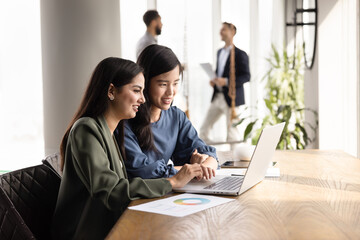 Two positive diverse female colleagues working on Internet marketing strategy together, typing on...