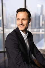 Positive handsome Latin businessman looking away with dreamy smile, posing at large office window...