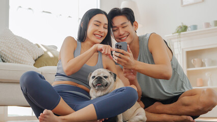 Asian couple checking smartphone together while sitting after workout, relaxing with pet dog at home