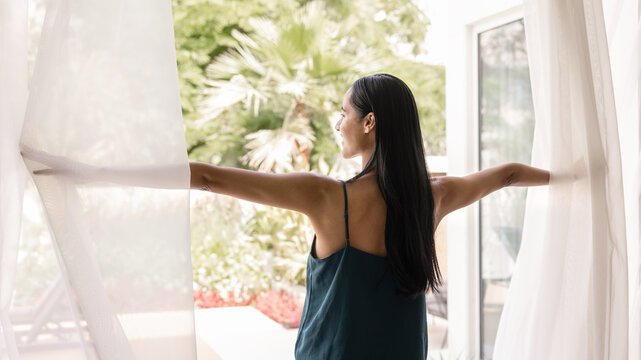 Happy young Maori woman opening veils at window or glass door, looking at terrace view, tropical greenery, enjoying vacation, holiday, weekend in hotel on modern resort. Back view, banner shot