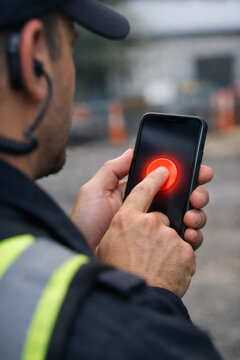 A security guard presses a digital red button on his smartphone, signaling alert or emergency response at an industrial site, a moment of readiness.