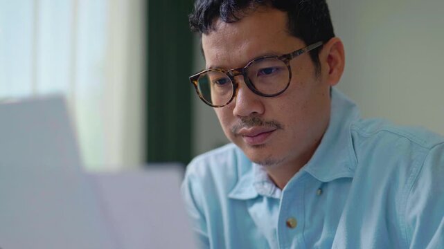 Close-up of Stressed Asian Man Holding Head in Pain While Checking Bills.