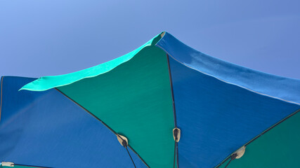 Green and blue beach umbrella. Blue sky in the background. View from below. Relaxing context. Summer holidays by the sea. General contest and location