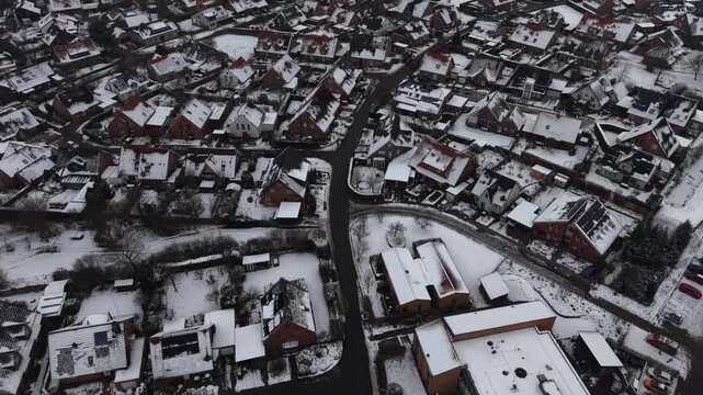 Top-down aerial flight over snow-covered residential neighborhood, showing rooftops, narrow streets and compact housing patterns during winter. Cleared Main Street of neighborhood in German town.