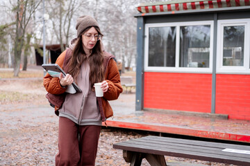 Young female freelancer in brown jacket and hat working remotely outdoors in autumn park, holding laptop and coffee.