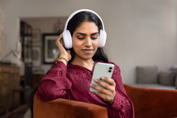 Happy young adult Indian girl in big headphones listening to music from online service, using...