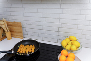 Chicken skewers cooking in a frying pan on an induction hob, surrounded by kitchen utensils, lemons, and oranges