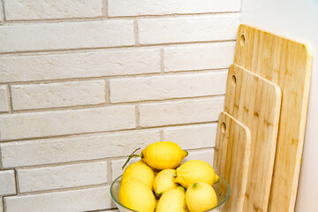 Fresh lemons in a clear glass bowl sitting on a counter next to bamboo cutting boards against a white brick wall