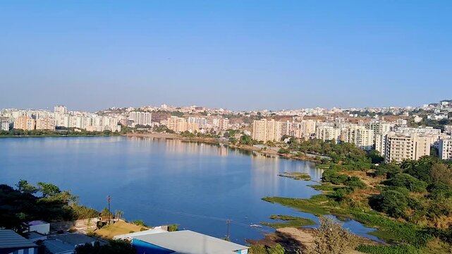 Urban riverside landscape with a calm water body, green vegetation in the foreground, and a modern residential skyline under a clear blue sky, showcasing peaceful city life and natural surroundings.