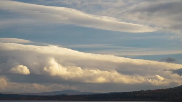 A wide shot captures dramatic, smooth, lens-shaped lenticular clouds layered above billowing cumulus clouds, creating a unique atmospheric display over a distant mountain range and forested landscape.