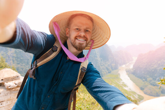 Smiling caucasian male tourist in traditional hat enjoying scenic ninh binh landscape. Travel Selfie photo concept