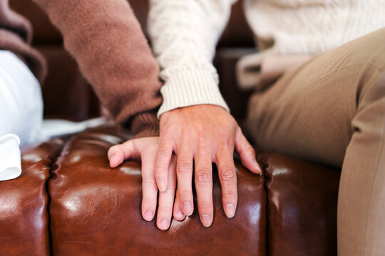 Two people sit closely on a brown leather couch, one gently placing their hand over the other&rsquo;s, capturing a tender moment of emotional support, comfort, and connection in a cozy indoor space.