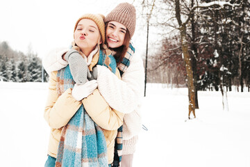 Fototapeta premium Two young beautiful smiling hipster female in trendy warm clothes and scarfs.Carefree women posing in the street in park. Positive pure models having fun in snow. Enjoying winter moments. Xmas concept