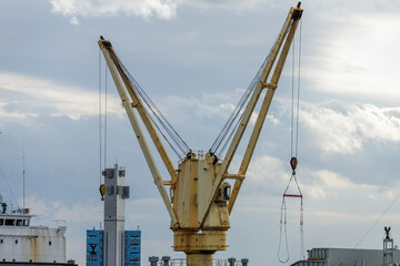 Landscape view of shipboard deck crane preparing to lift heavy loads with cables and hanging hook dangling in midair against cloudy sky backdrop over a shipyard.