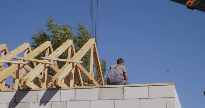 Young male builder in overalls sitting on a concrete block wall and hammering nails into wooden rafters while constructing the roof frame of a new residential house on a sunny day.