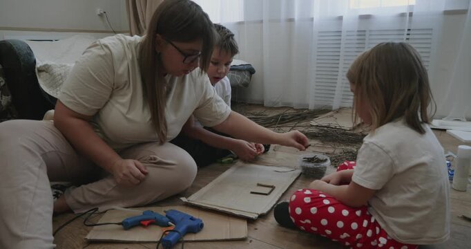 Caring mother with her son and daughter sitting on the floor at home, creating crafts from natural materials like twigs and using a hot glue gun for a fun family arts project.