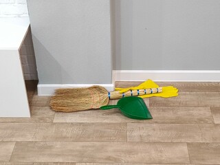 Straw broom, green dustpan, and yellow rubber gloves on a clean wooden floor with baseboard. Home...