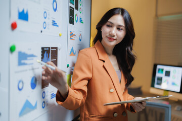 Asian businesswoman analyzing and presenting financial data on a whiteboard, using papers with various charts and graphs to illustrate business statistics and planning strategies
